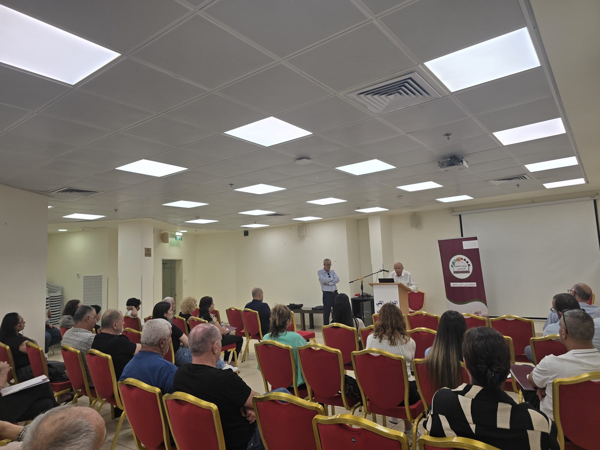 Seated audience in rows facing a speaker at the podium, with the SDC banner behind — Arab Civic Council founding assembly, Haifa, 1 July 2026.